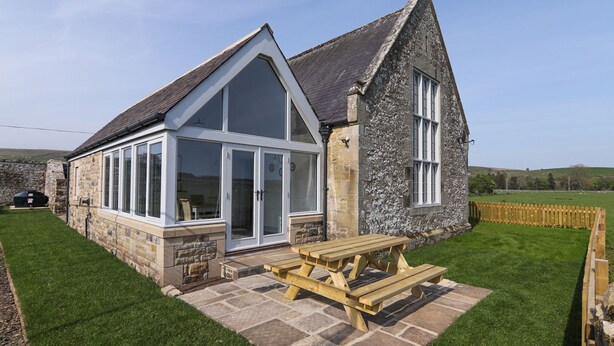 Une maison en pierre avec une grande fenêtre en verre et une table de pique-nique en bois dans le jardin.