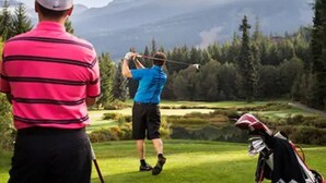 Un campo de golf con montañas al fondo, un jugador con camisa azul preparándose para golpear la pelota, y un bolso de golf en primer plano.
