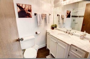 Ein Badezimmer mit einer weißen Toilette, einem Doppelwaschbecken mit großem Spiegel und zwei Handtüchern an einem Haken.