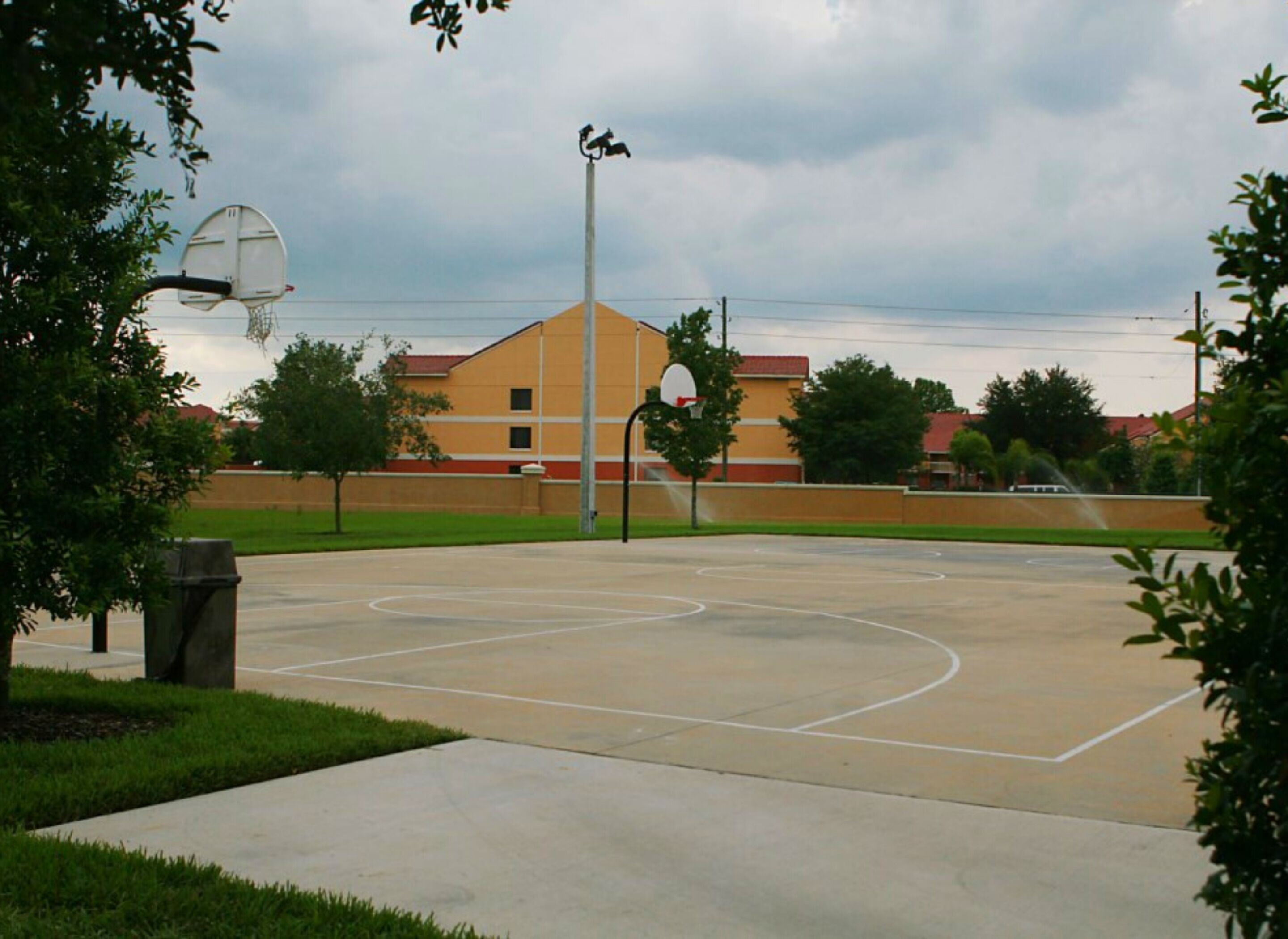 An outdoor basketball court with a hoop, a trash can, and a building in the background.
