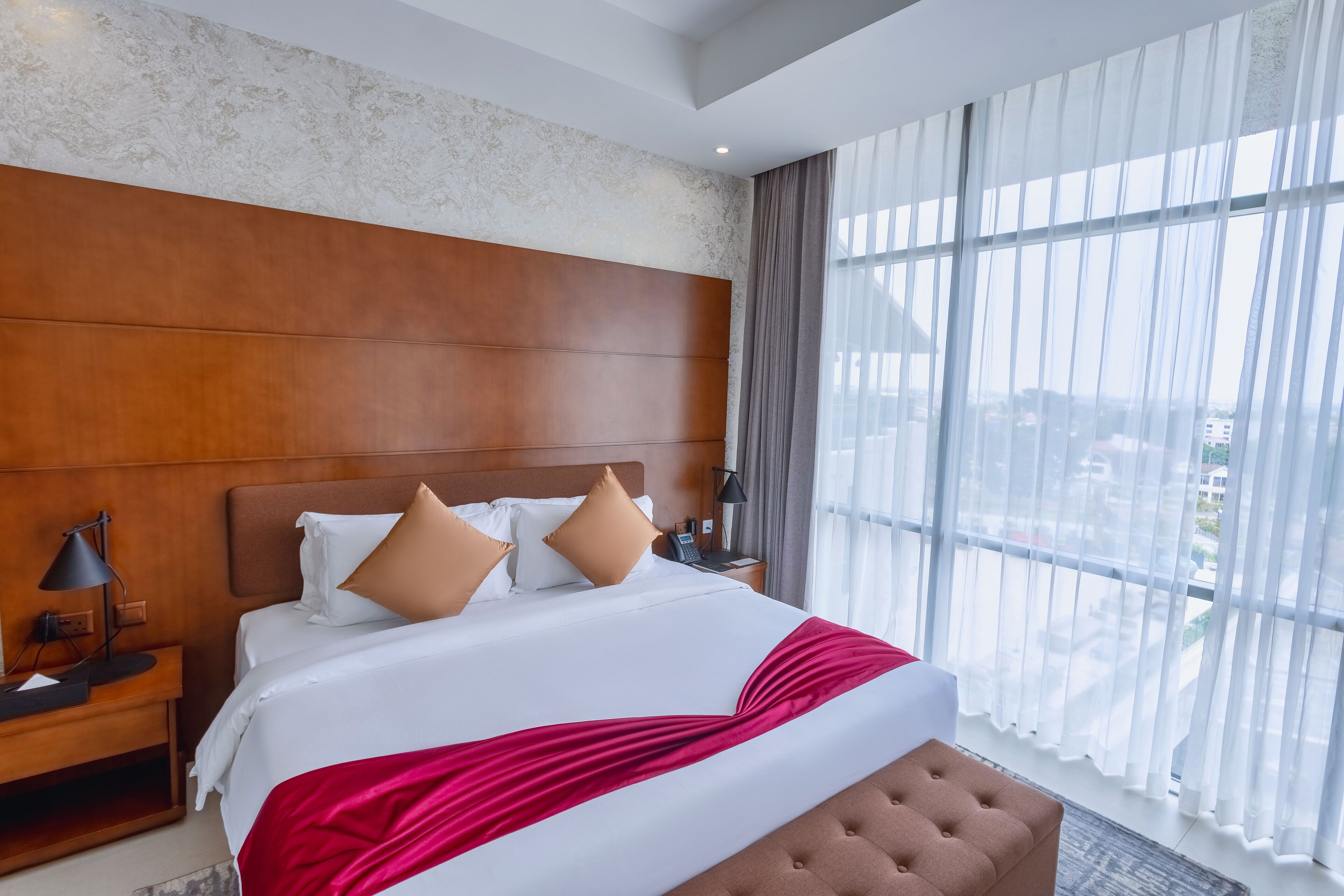 Habitación de hotel con una cama grande, cabecera de madera, mesita de noche con lámpara y vistas a la ciudad a través de amplias ventanas.