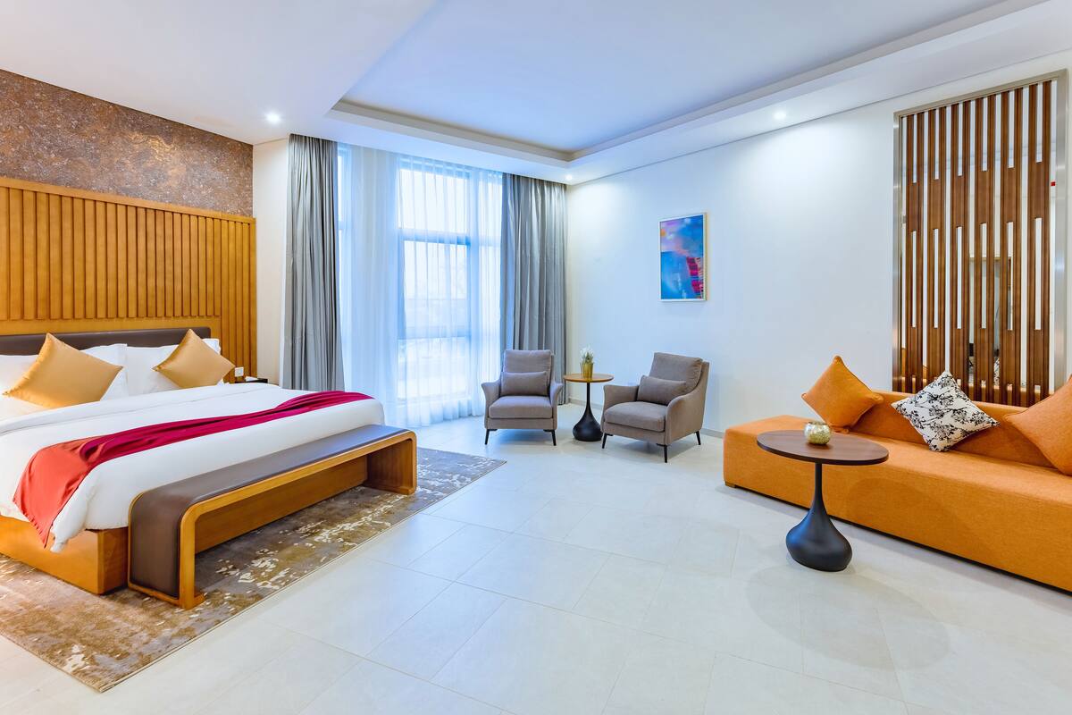 Ein modernes Hotelzimmer mit einem großen Bett, einer Couch, zwei Sesseln, einem kleinen Tisch und einem großen Fenster mit Vorhängen.