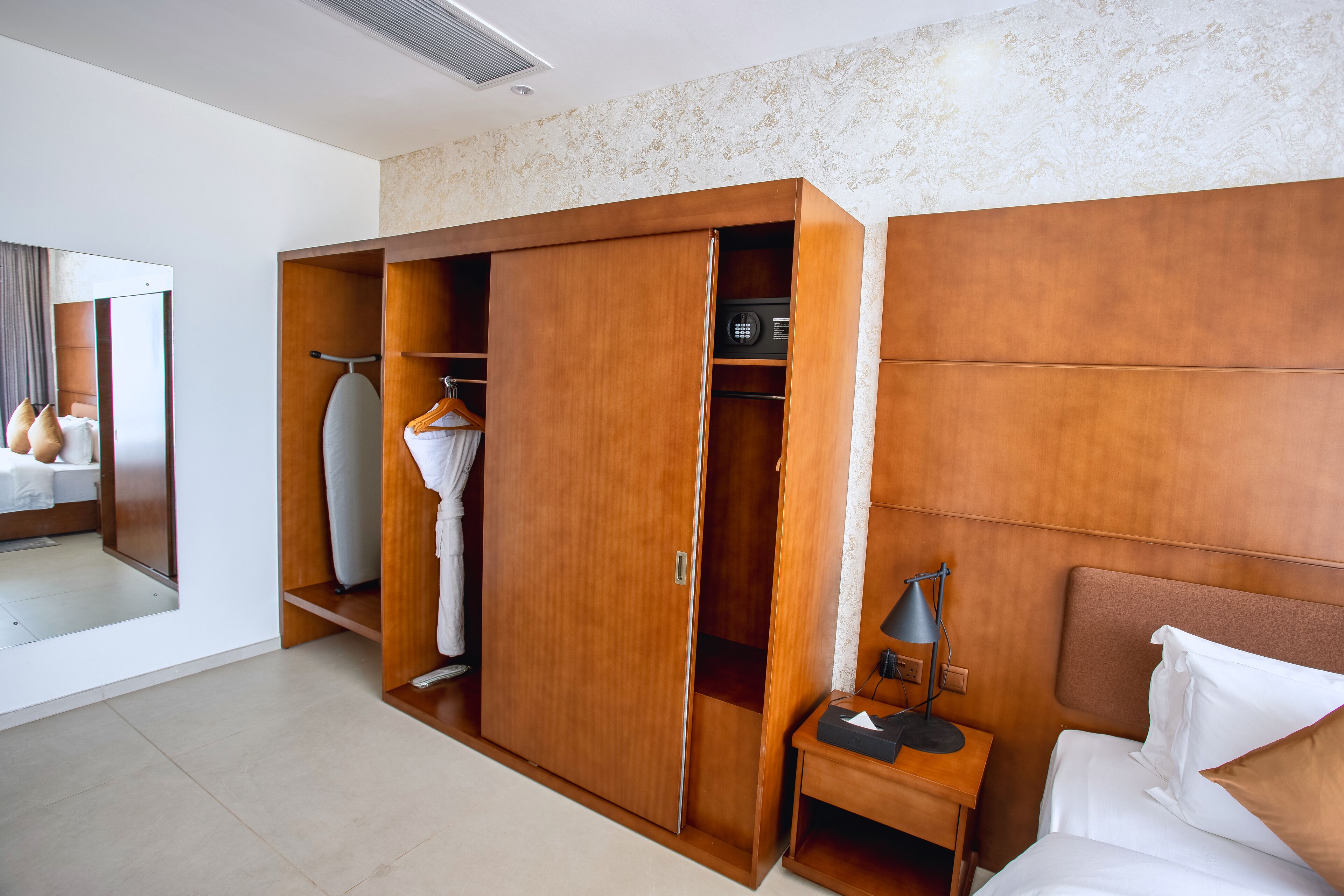 A hotel room with a large wardrobe, a bed, a bedside table with a lamp, and a mirror.