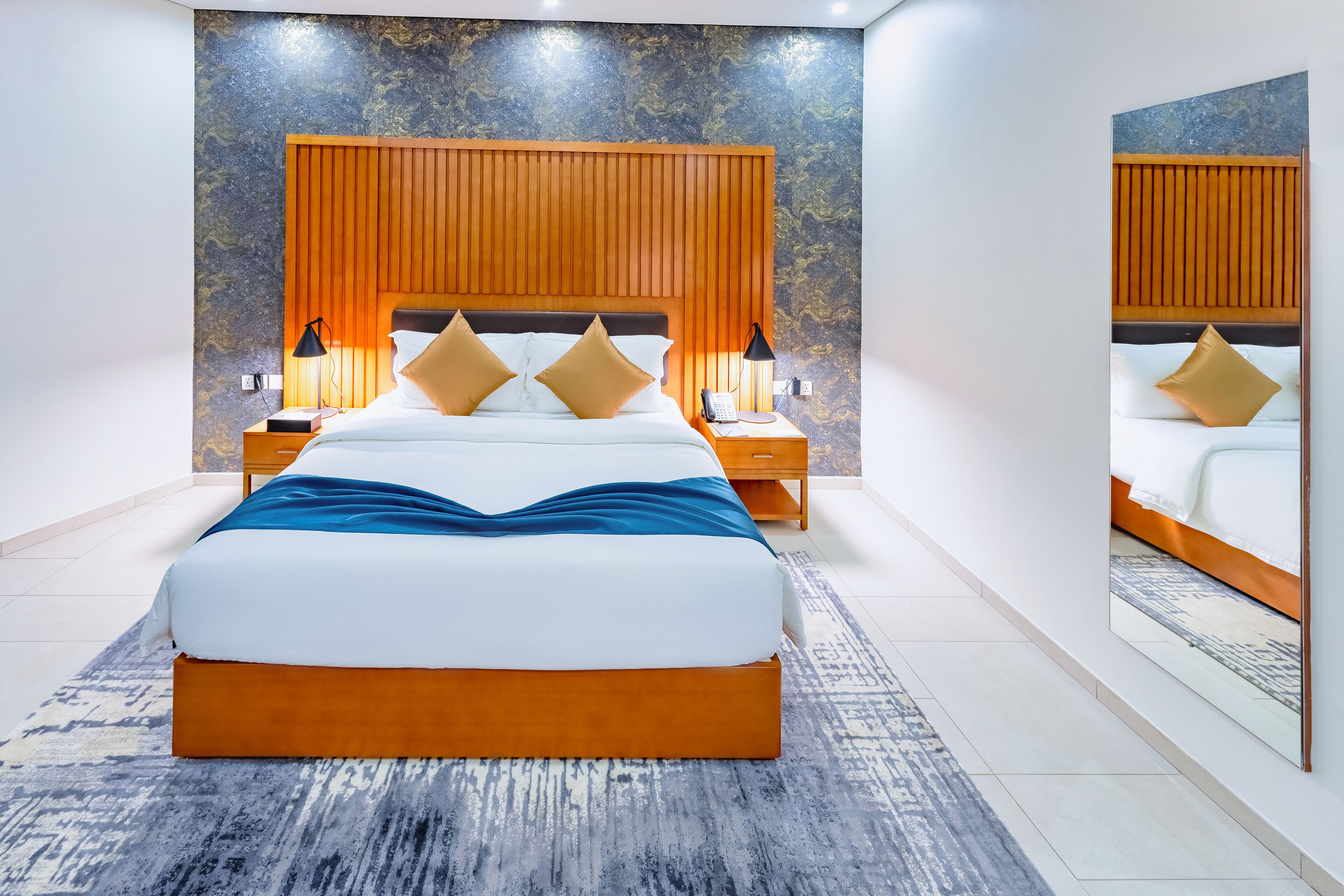 A modern bedroom with a large bed, wooden headboard, two bedside tables with lamps, and a patterned rug.