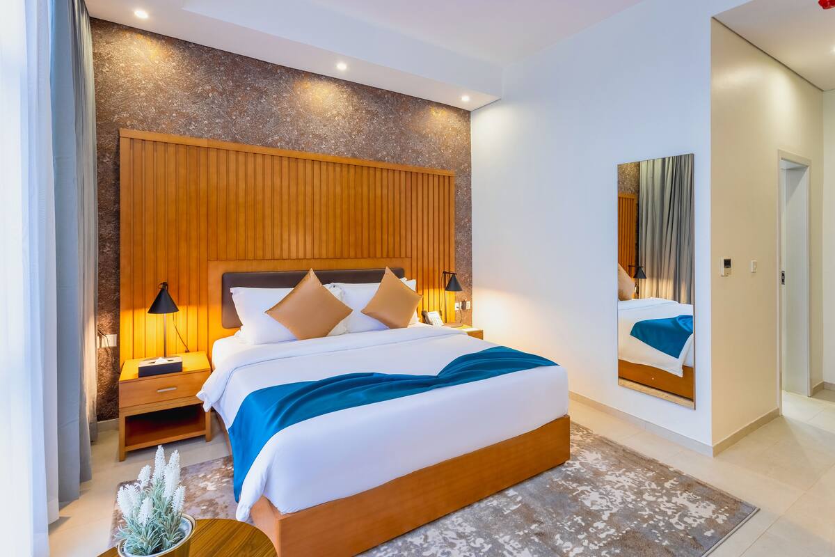 Ein modernes Schlafzimmer mit einem Holzkopfende, einem großen Bett mit weißer und blauer Bettwäsche, zwei Nachttischlampen und einer kleinen Pflanze auf dem Nachttisch.
