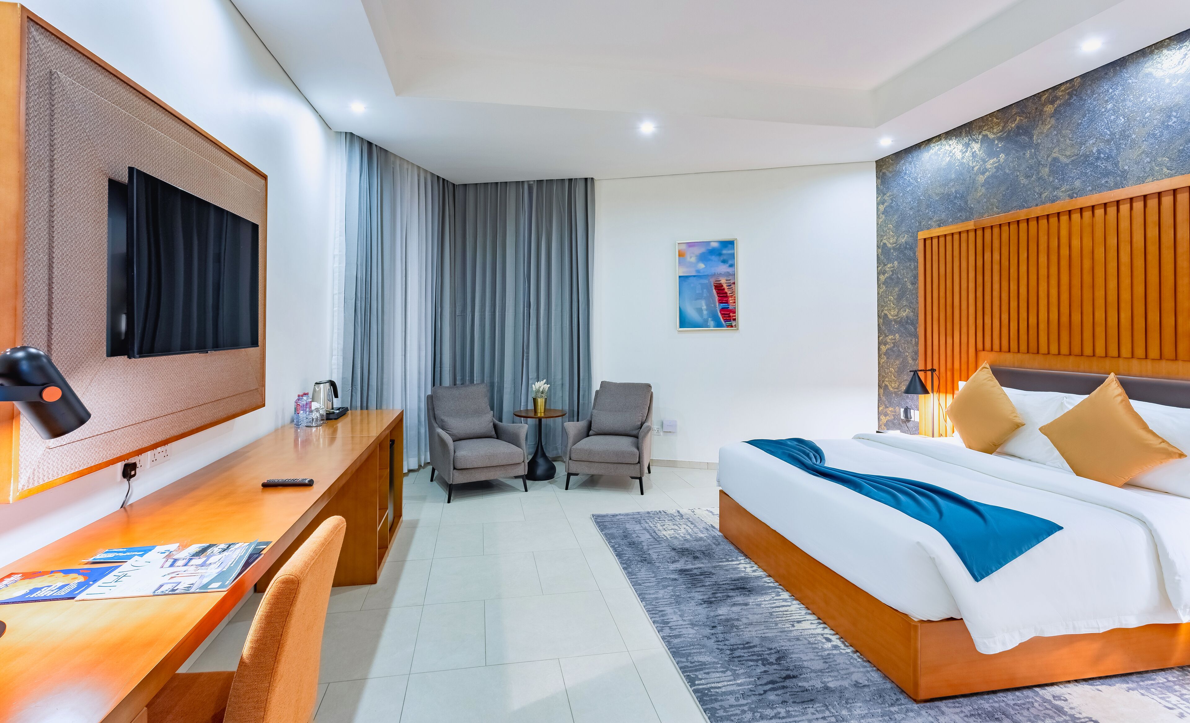A hotel room with a bed, a desk, two armchairs, a TV, and a painting on the wall.