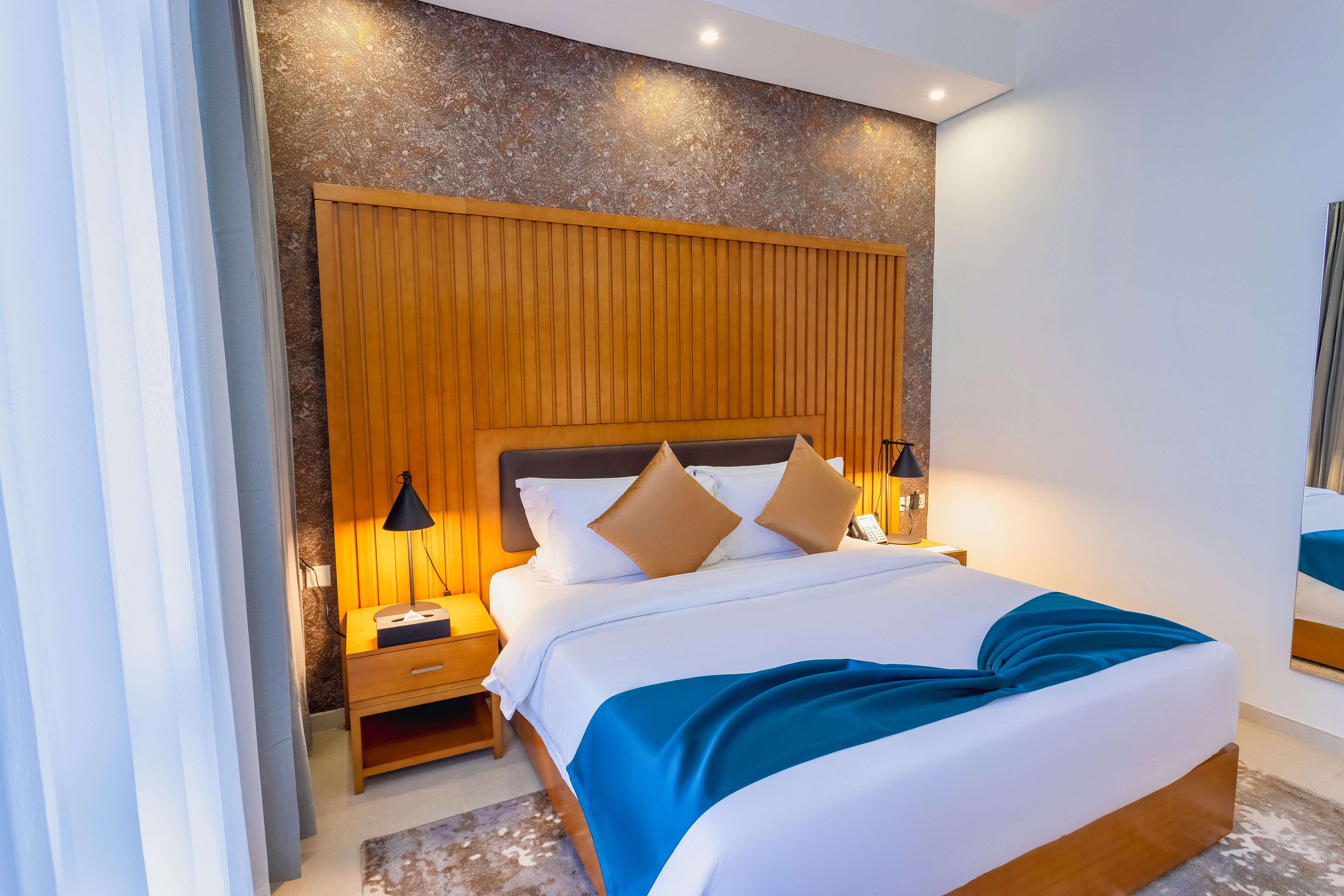 Una habitación de hotel moderna con cabecera de madera, una cama grande con ropa de cama blanca y azul, dos mesitas de noche con lámparas y una pared texturizada detrás de la cama.
