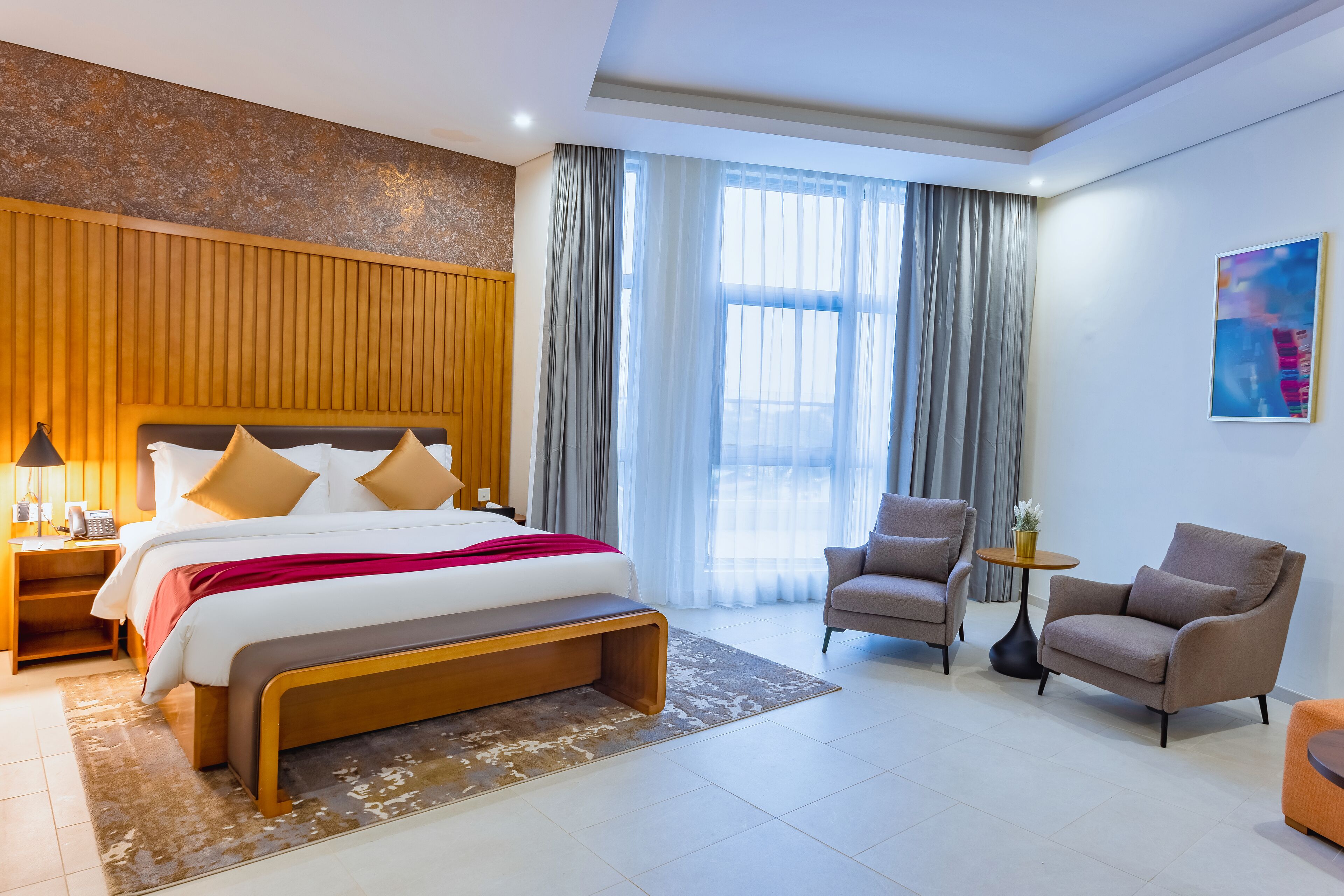 A modern hotel room with a large bed, a bench, two armchairs, a small table, and a painting on the wall.