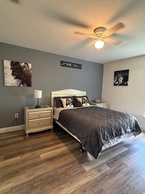 A bedroom with a bed, nightstand, lamp, ceiling fan, and artwork on the walls.