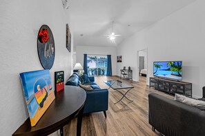 Un salon moderne avec un canapé, une table basse et un téléviseur. Un tableau orne le mur et un panneau d’accueil est visible.