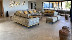 Amplia sala de estar con un sofá modular grande, un sofá más pequeño y dos otomanas. Hay una mesa de comedor con sillas y un ventanal con vistas al exterior.