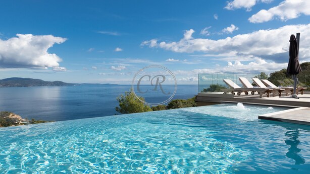 Piscina azul cristalina con sillas de descanso y una sombrilla, con vistas a una extensión de agua y tierra a lo lejos.