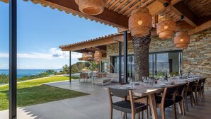 Un área de comedor al aire libre con vista al océano, paredes de piedra y lámparas de fibra.