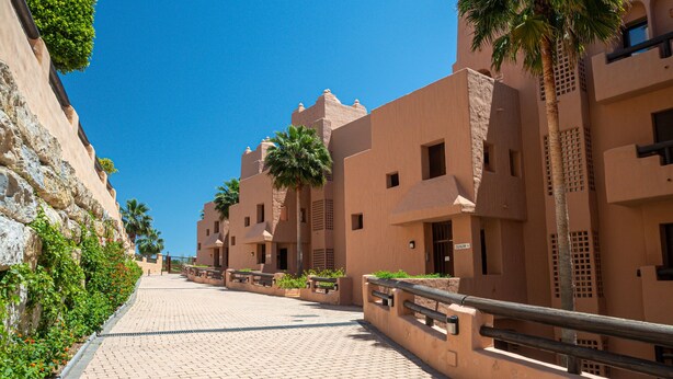 A pathway lined with buildings featuring a uniform architectural style, a clear sky, and palm trees. - Coto Real 2359 Lovely apartment with sea view (Manilva)