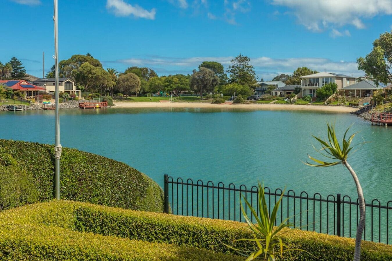 A serene lakeside view with a well-maintained hedge, a metal fence, and a clear sky.