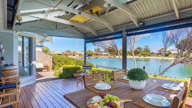 Uma área de refeições ao ar livre com uma mesa de madeira posta para uma refeição, com vista para um lago e um jardim paisagístico.