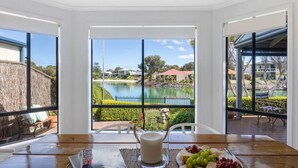 Uma área de refeições com uma mesa posta para dois, um jarro de leite e um prato de frutas. Uma grande janela oferece vista para um lago e edifícios ao redor.
