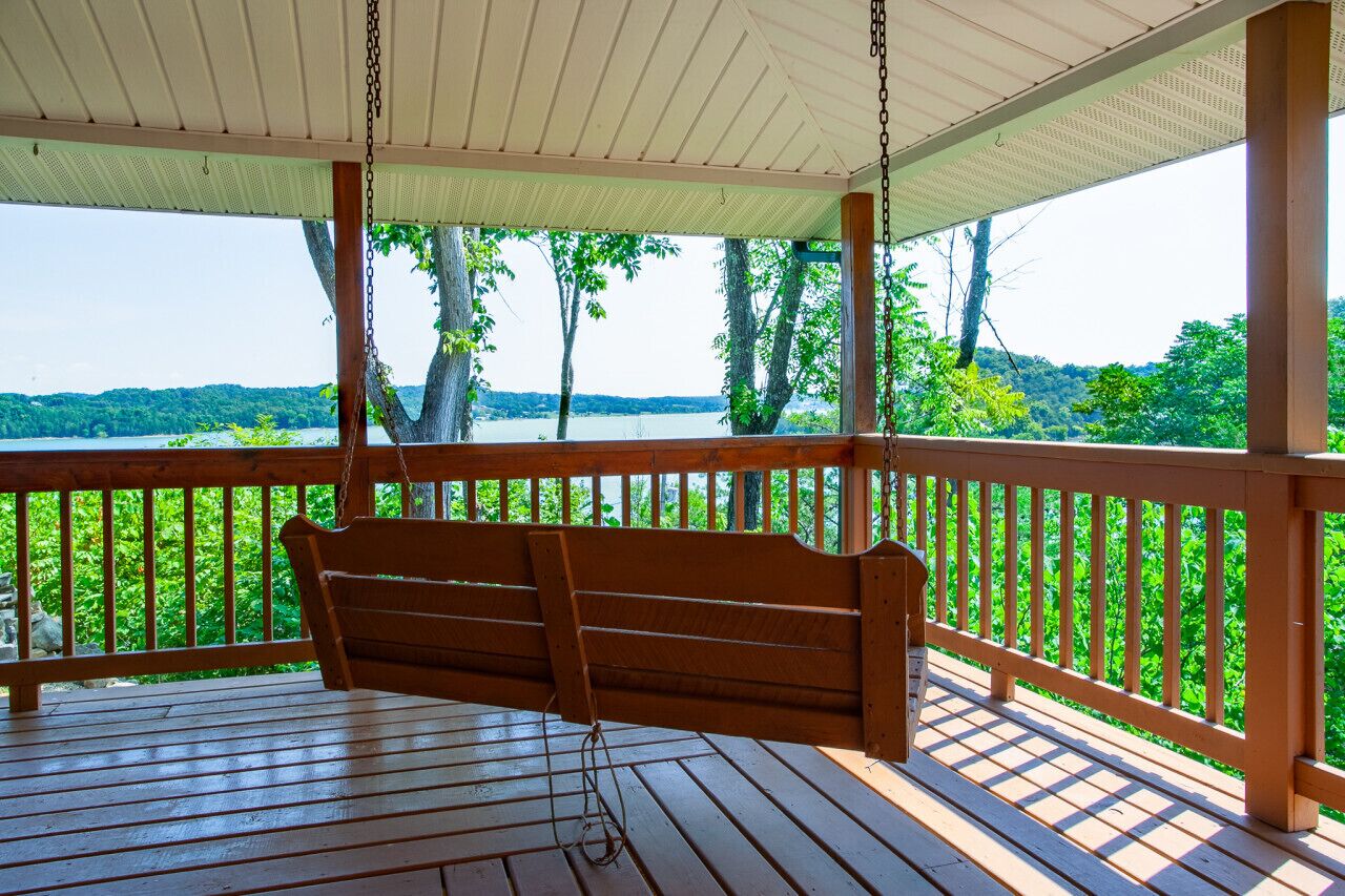 A wooden swing on a covered porch overlooking a lake with trees.