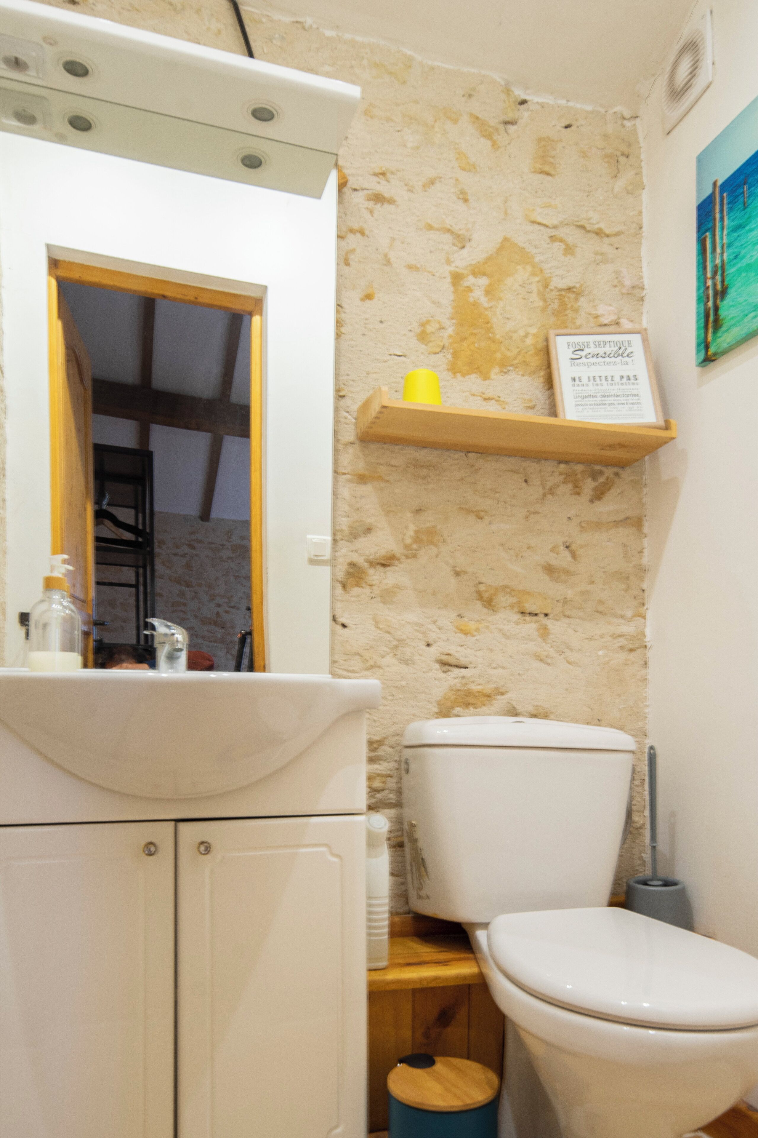 Een badkamer met een wit wastafel, toilet, houten plankje met een geel beker, en een bord aan de muur.