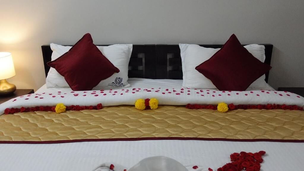 Un lit bien fait, avec une tête de lit matelassée, deux oreillers rouges et des pétales de rose éparpillés.