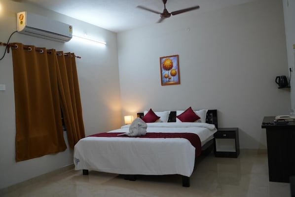 A bedroom with a bed, a nightstand, a desk, a wall-mounted air conditioner, and a ceiling fan. - Hotel Akshith Inn (Thanjavur)