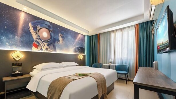 A hotel room with a bed, a desk, a chair, a nightstand, a wall-mounted TV, and a mural of an astronaut. - Jiangmen Danxia Hotel (Jiangmen)