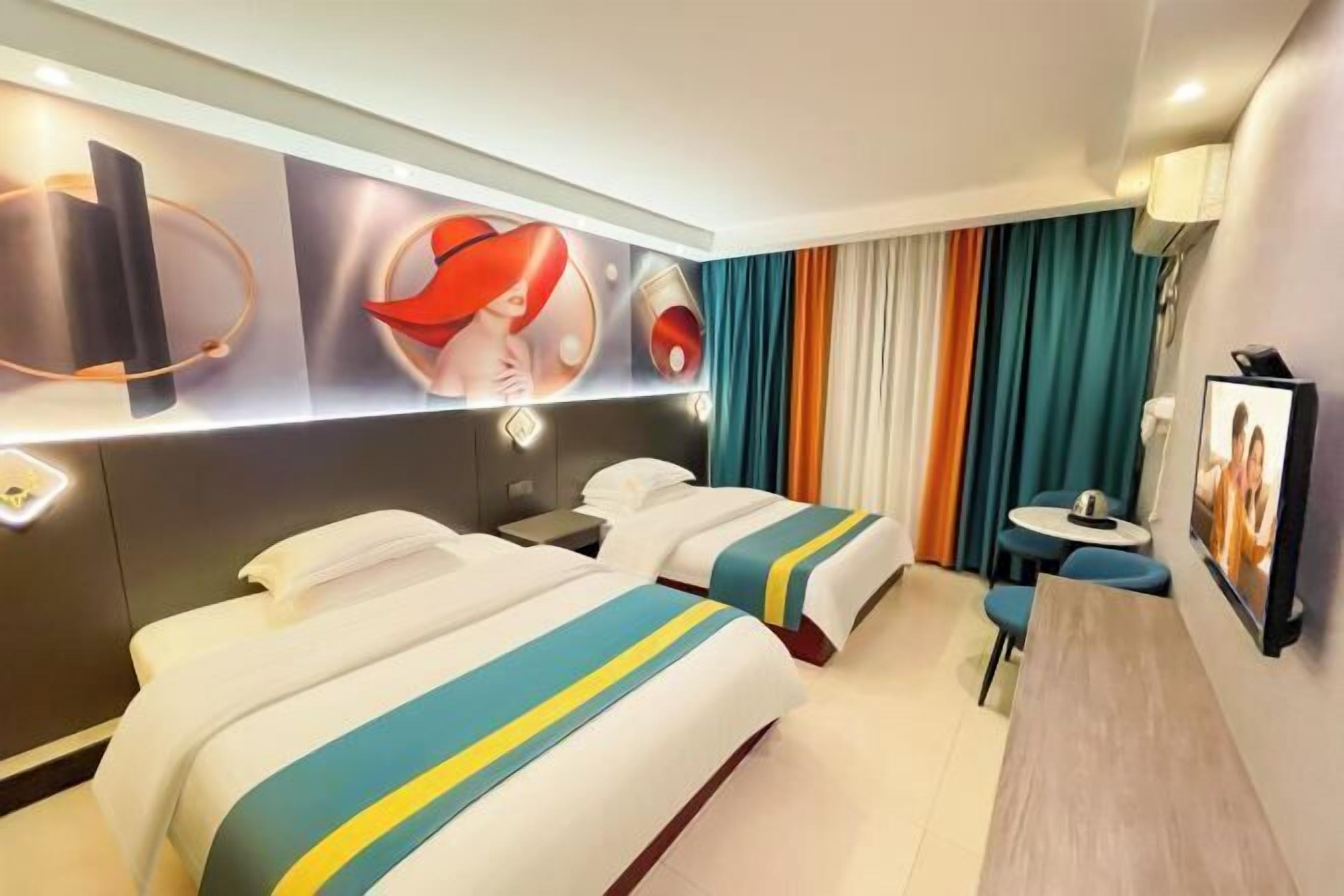A hotel room with two beds, a small table, a chair, a desk, a lamp, and a wall-mounted TV.