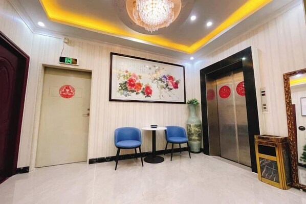 A hotel lobby with two blue chairs, a round table, a chandelier, an elevator, and a framed artwork on the wall. - Jiangmen Danxia Hotel (Jiangmen)