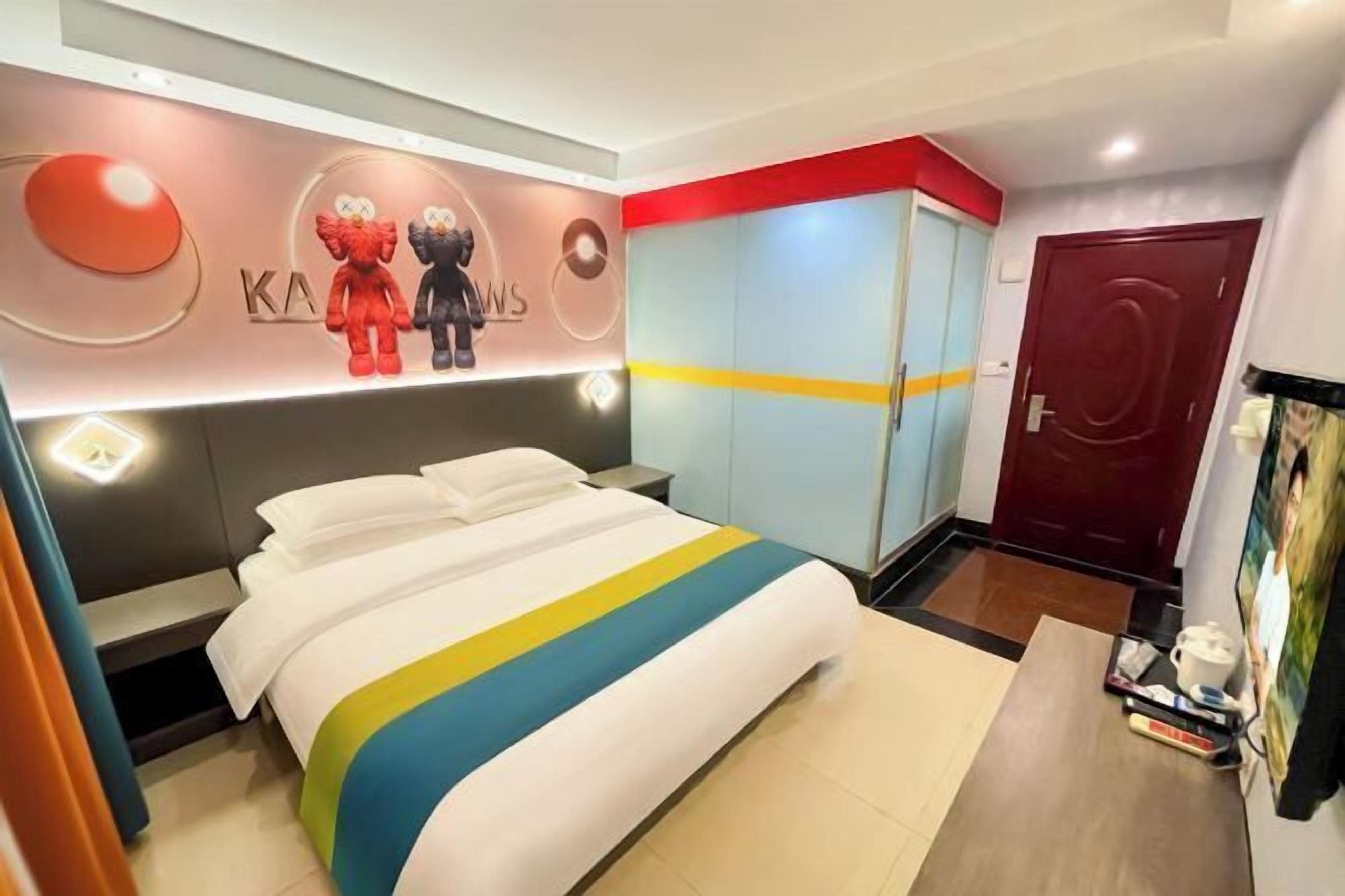 A modern hotel room with a bed, a wardrobe, a red door, and a wall with a logo.