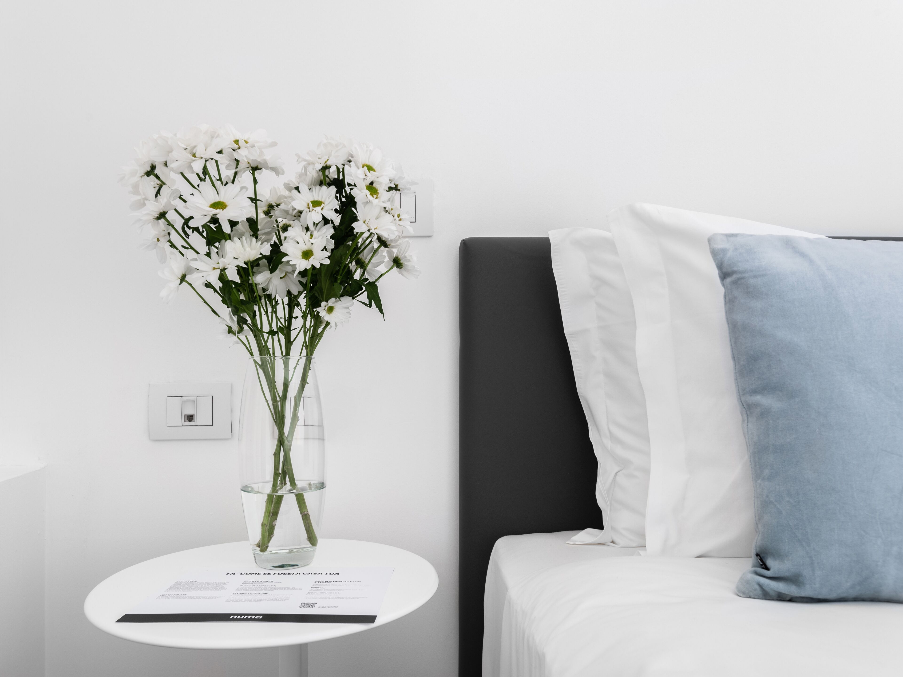 A modern hotel room with a white bed, a blue pillow, a small white table with a vase of white flowers, and a white wall with a light switch.