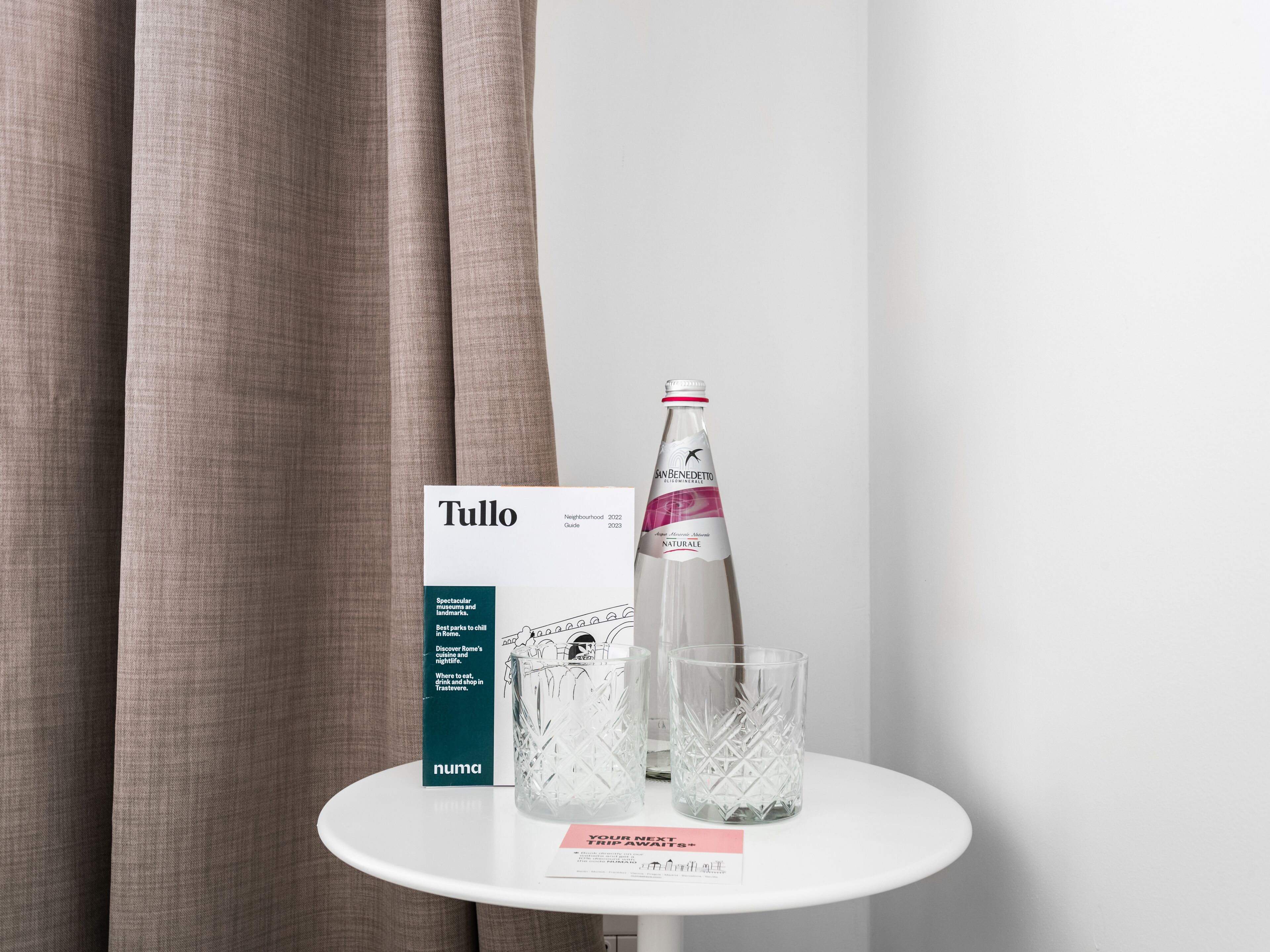 A table with a bottle of San Benedetto sparkling water, two crystal glasses, a Tullio neighborhood guide, and a 'Your Next Trip Awaits' card.