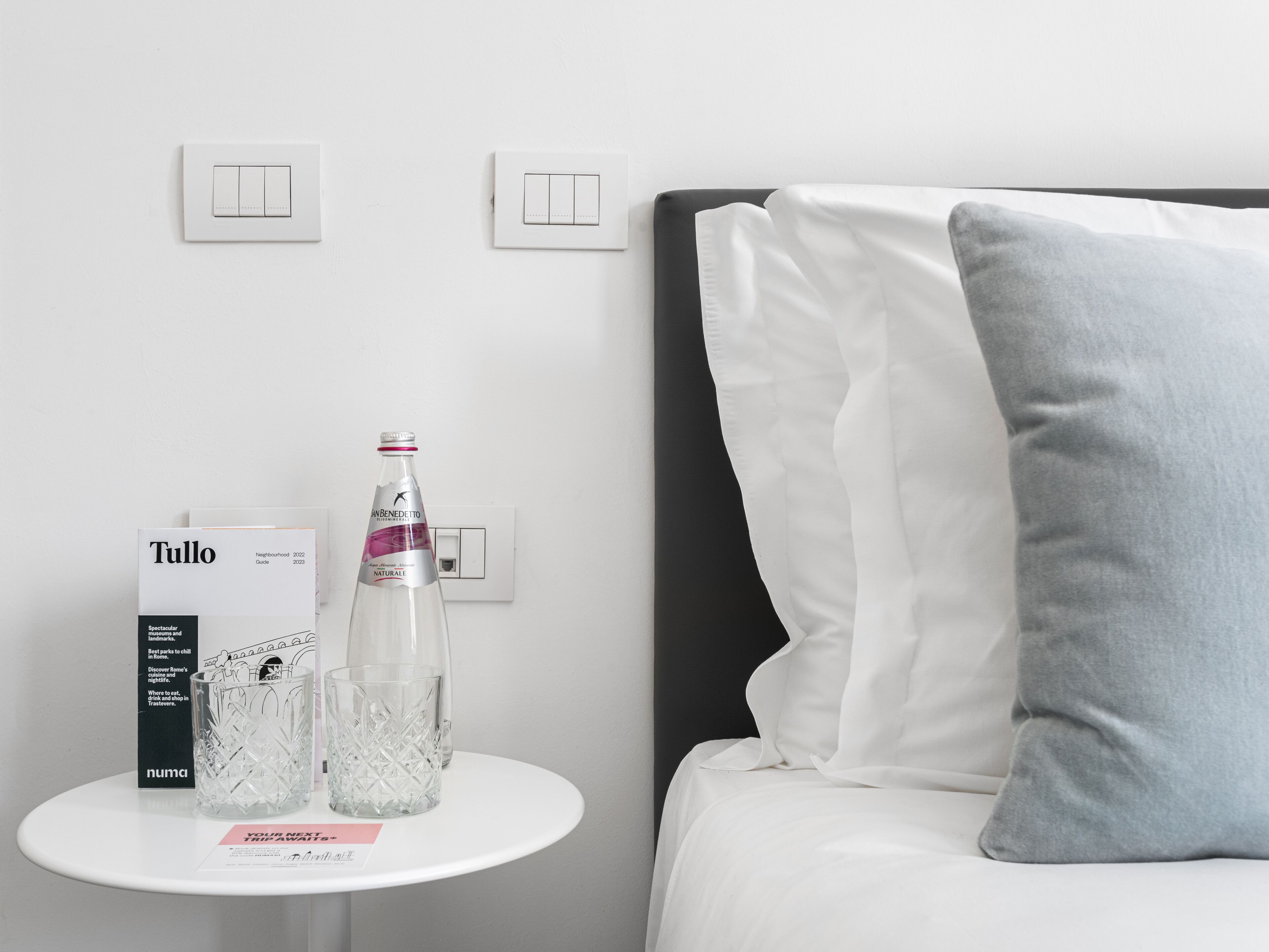 A hotel room with a white bed, a grey pillow, a small white table with a bottle of water, a glass, and a brochure.