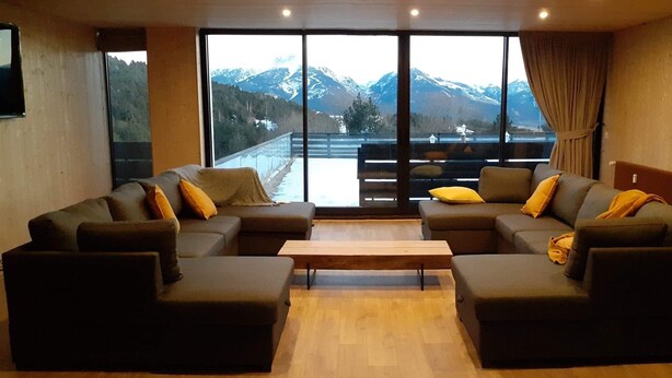 A living room with two sofas and a coffee table, large windows with a mountain view, and a balcony with a railing.
