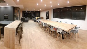 A modern kitchen with dark cabinetry, a central island, and a dining area with a long wooden table and patterned chairs.