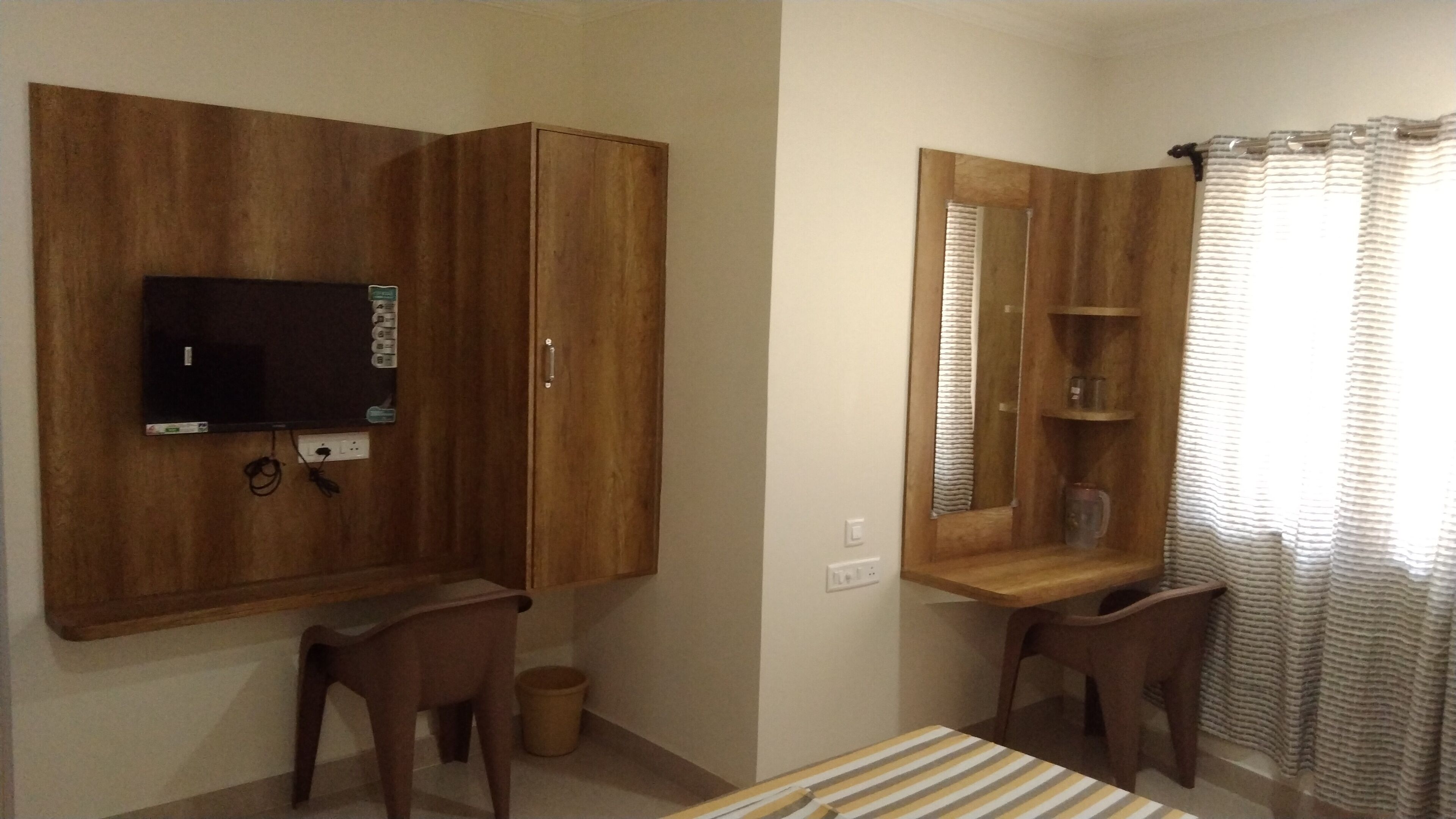 A bedroom with a wooden wardrobe, a television mounted on the wall, a small table, and a mirror with shelves.