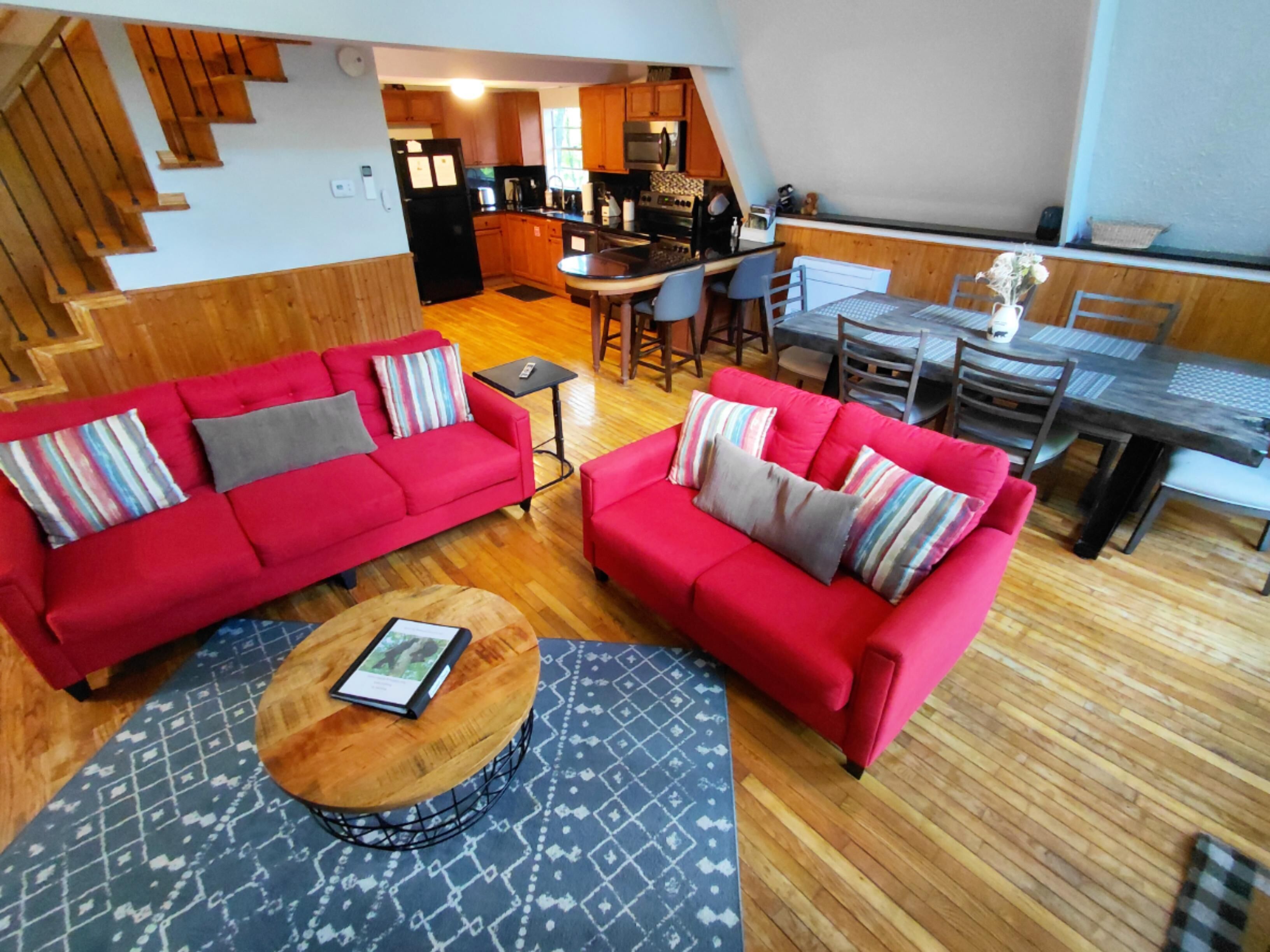 Un vaste espace de vie comprenant un canapé rouge, une table basse ronde et une table à manger avec des chaises. On aperçoit à l’arrière-plan une cuisine avec des meubles en bois et un coin repas avec une table haute et des chaises.