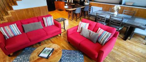 Un vaste espace de vie comprenant un canapé rouge, une table basse ronde et une table à manger avec des chaises. On aperçoit à l’arrière-plan une cuisine avec des meubles en bois et un coin repas avec une table haute et des chaises.