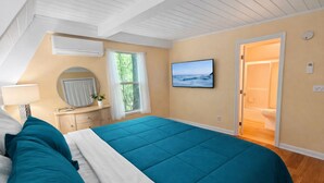 A bedroom with a large bed, a TV, a window with curtains, a bedside table with a lamp, and a door leading to a bathroom.