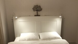 Un lit avec une tête de lit blanche et deux oreillers, une petite décoration en forme d’arbre accrochée au mur et une table de chevet avec une lampe.