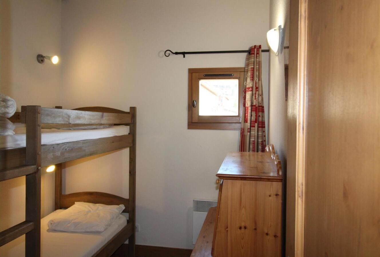 A small, cozy room with bunk beds, a wooden dresser, a window with a view, and a wall-mounted light fixture.
