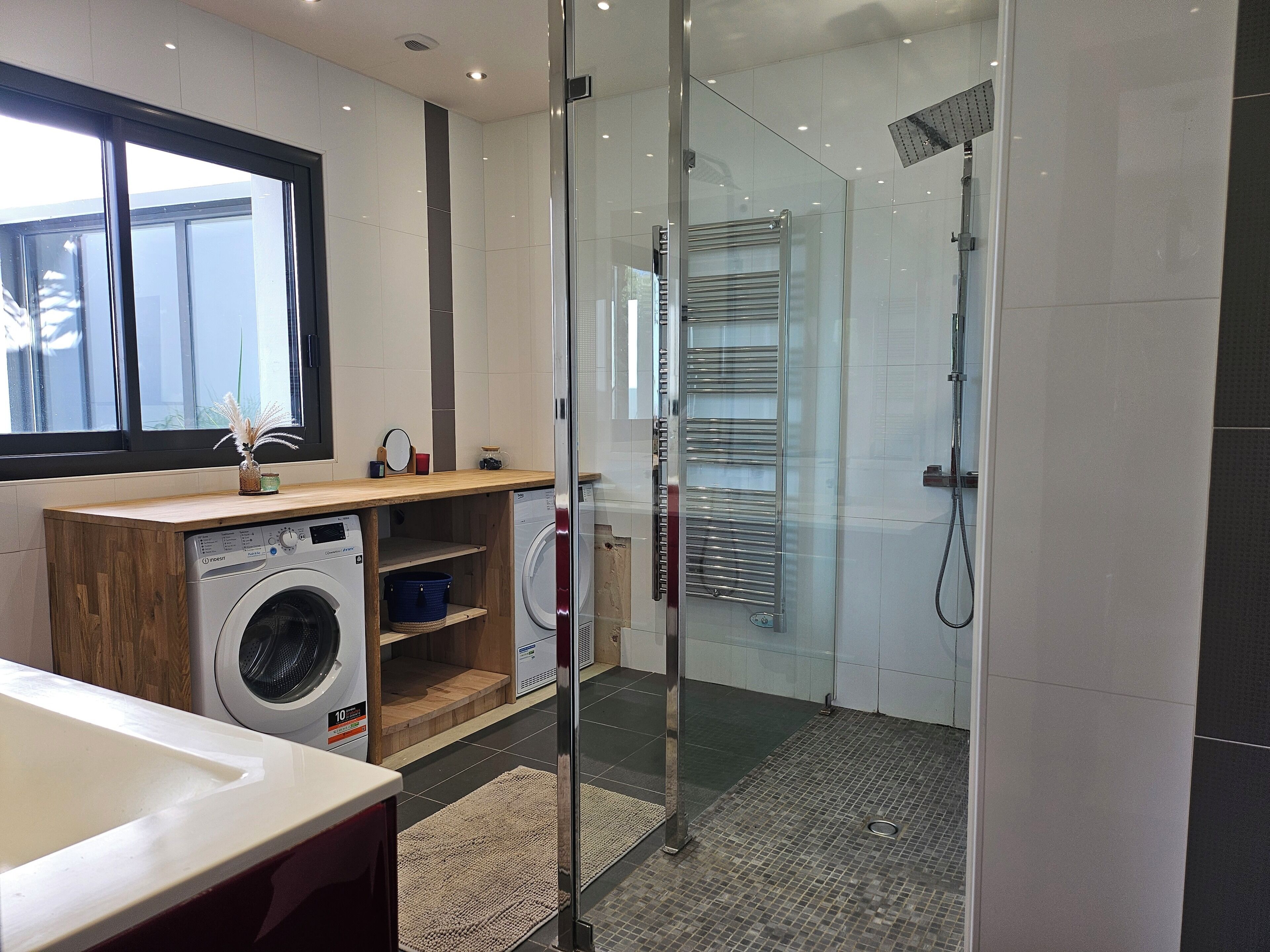 A modern bathroom with a glass shower enclosure, a built-in washing machine, and a wooden vanity.