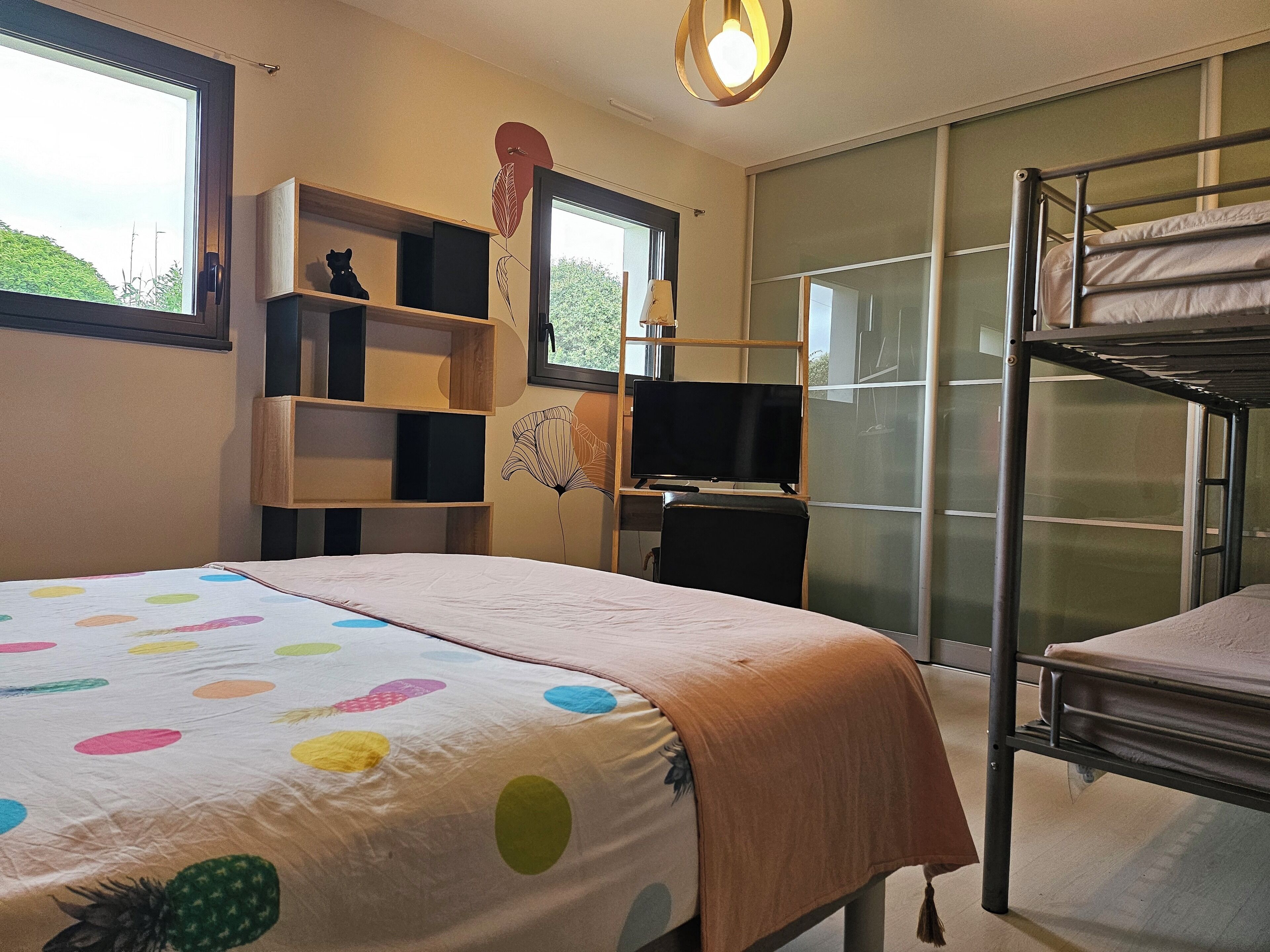A bedroom with a bunk bed, a desk with a computer, a TV, and a window with a view of greenery.