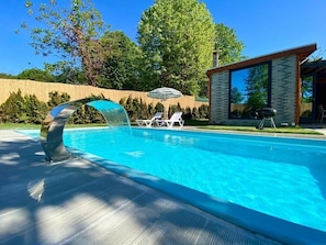 A modern swimming pool with a unique arch structure, surrounded by a wooden fence and lush greenery.