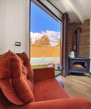 A modern living room with a large window, a red sofa, a small table with a kettle, and a wood-burning stove.