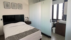 Et moderne soveværelse med seng, ventilator, sengebord og et badevrelse, der kan ses gennem et glasafskærmet område.