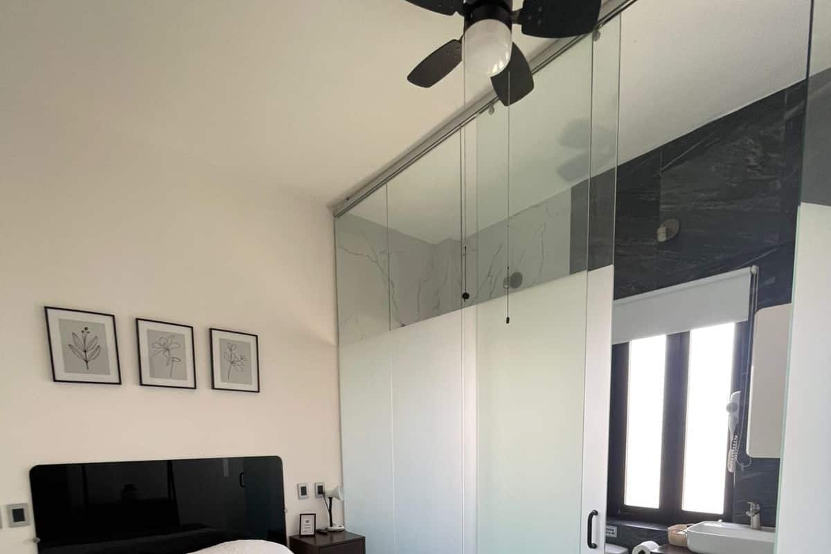 A modern bedroom with a bed, a ceiling fan, a nightstand, and a bathroom visible through a glass partition.
