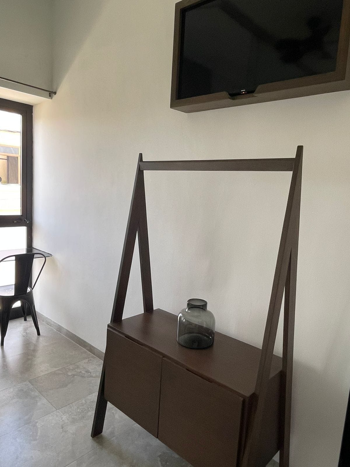 Una habitación con un escritorio y una silla de madera, un televisor montado en la pared y una ventana que proporciona luz natural.