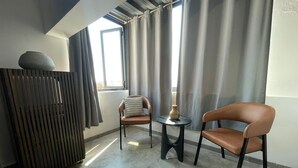 Et moderne værelse med højt til loftet, et stort vindue med gardiner, en sofa, en lænestol, et lille bord med en vase og en radiator.