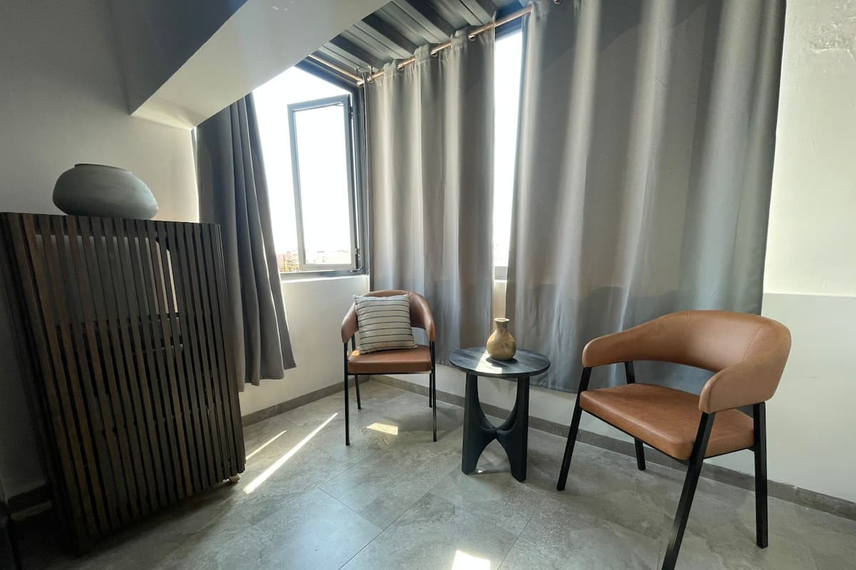 A modern room with a high ceiling, a large window with curtains, a sofa, a chair, a small table with a vase, and a radiator.