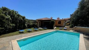 A rectangular swimming pool with clear blue water, surrounded by a stone deck. There are lounge chairs and a small building with a tiled roof in the background.